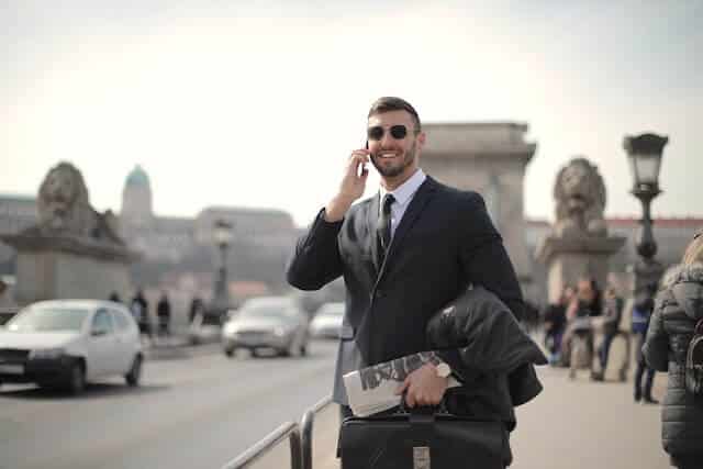 A man in formal attire, looking pleased, holds a smartphone and stands in a posh environment, indicating satisfaction with the Mynt Models escorting agency services.